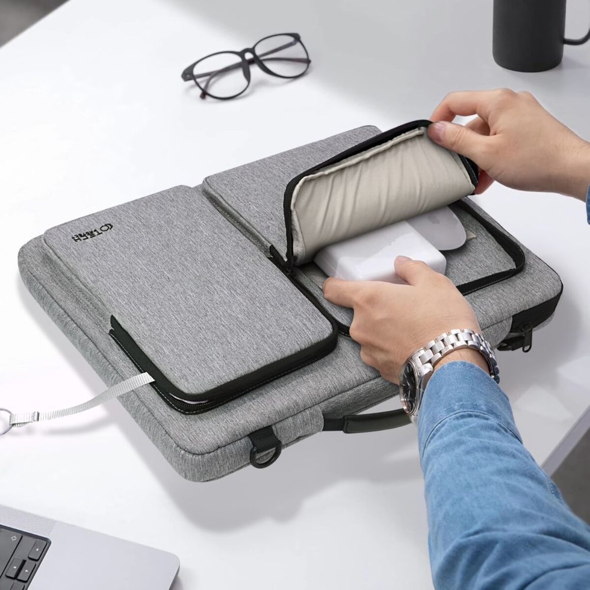 Graue Laptoptasche mit weißen Gegenständen, Brille und Tasse im Hintergrund.