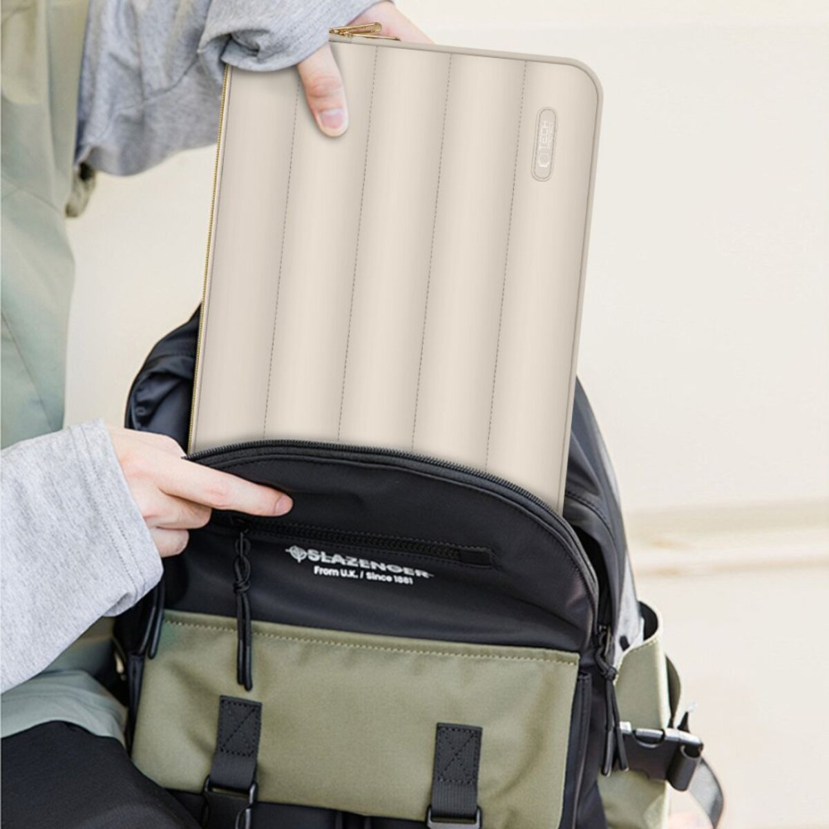 Hände stecken eine cremefarbene gepolsterte Hülle in einen schwarz-grünen Rucksack.