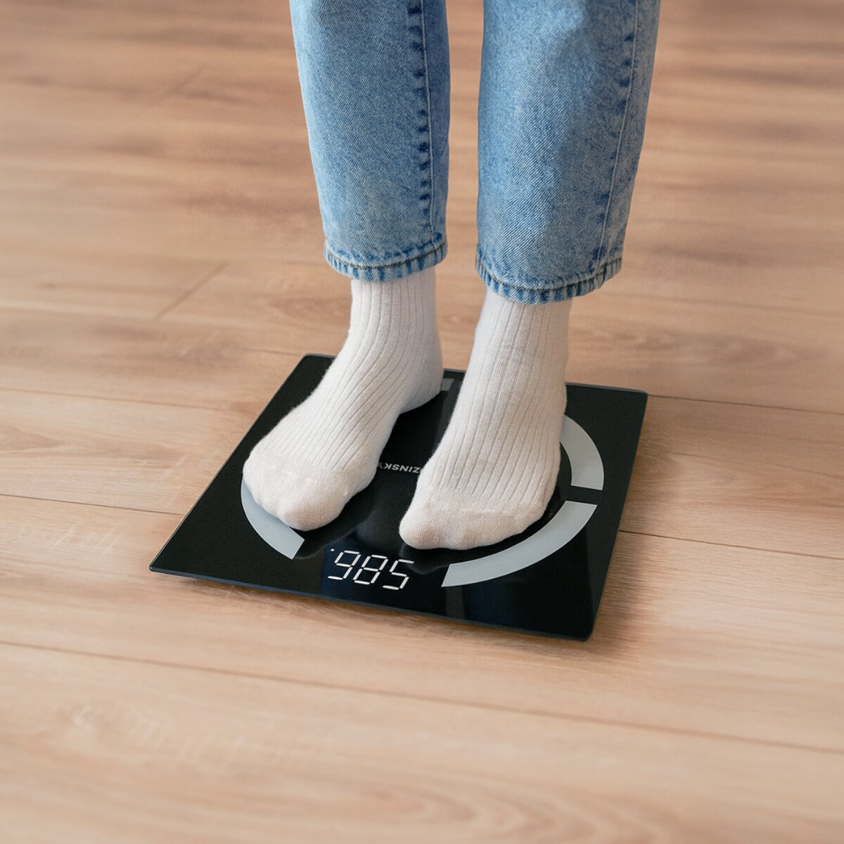 Person in Jeans und weißen Socken steht auf digitaler Waage mit Anzeige 98.5.