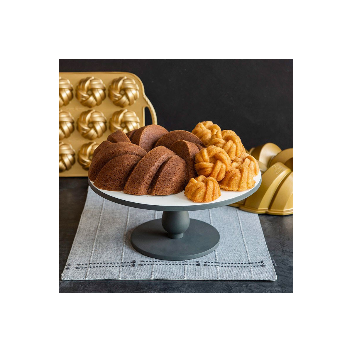 Beyaz bir kek standında çeşitli bundt kekler, bazıları koyu kahverengi, bazıları daha açık, arkada altın rengi bundt kalıpları bulunuyor.
