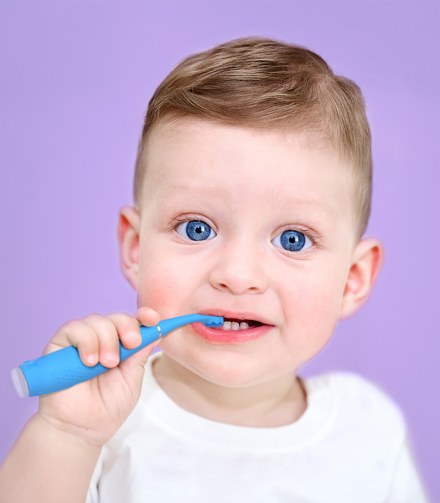 Un bambino con occhi azzurri brillanti e capelli castani, che indossa una maglietta bianca, si lava i denti con uno spazzolino blu.