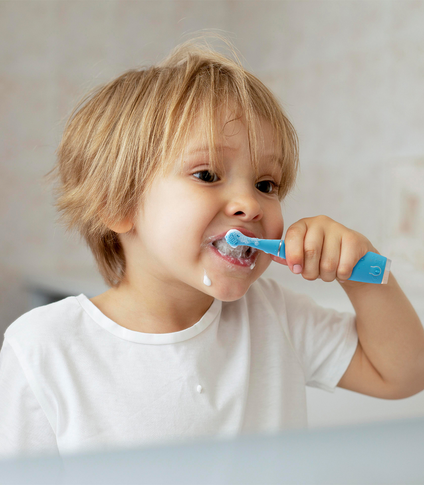 Un bambino biondo in maglietta bianca si lava i denti con uno spazzolino elettrico blu, schiuma intorno alla bocca.