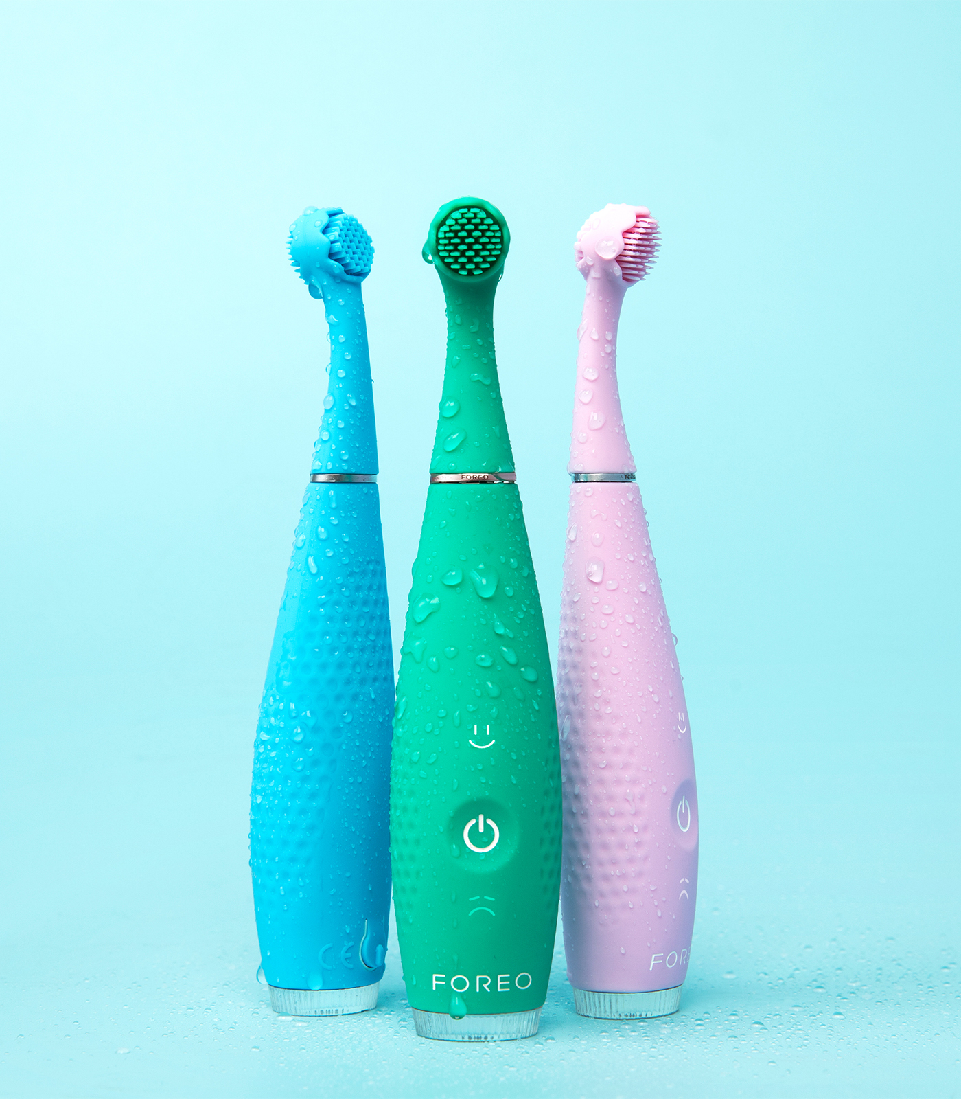 Tre spazzolini elettrici bagnati, blu, verde e rosa, in piedi su uno sfondo azzurro.