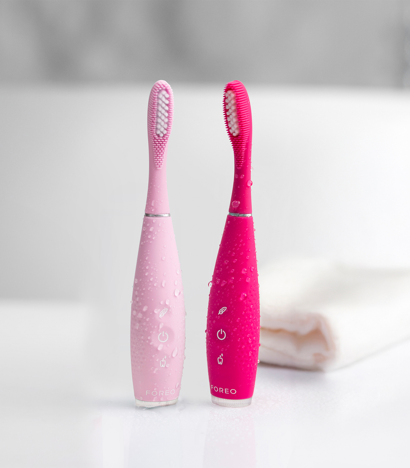 Due spazzolini elettrici bagnati, uno rosa chiaro e uno fucsia, sono in piedi su una superficie bianca con un asciugamano sullo sfondo.
