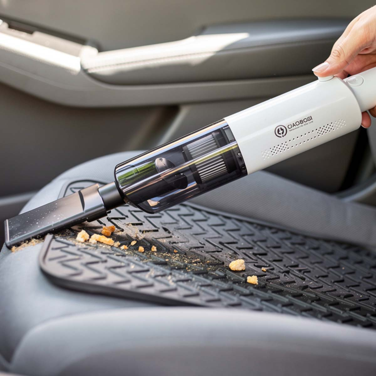 Un aspirapolvere bianco portatile viene usato per aspirare briciole da un tappetino nero per auto.