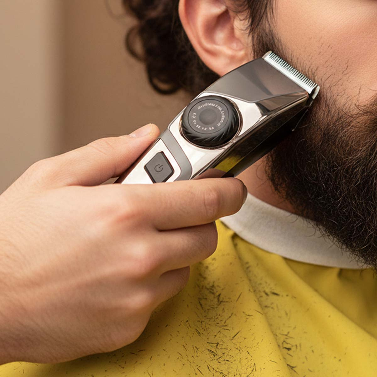 Una mano sostiene un recortador plateado para recortar una barba. Hay pelos en una capa amarilla.