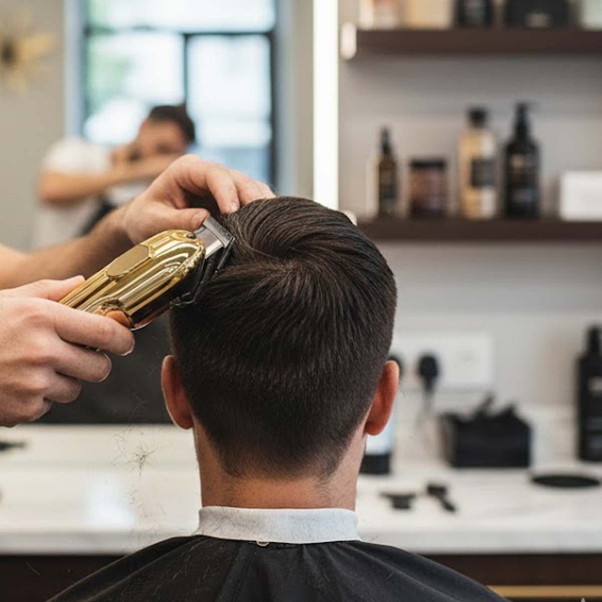 Coiffeur coupant les cheveux avec une tondeuse dorée, client portant une cape noire.