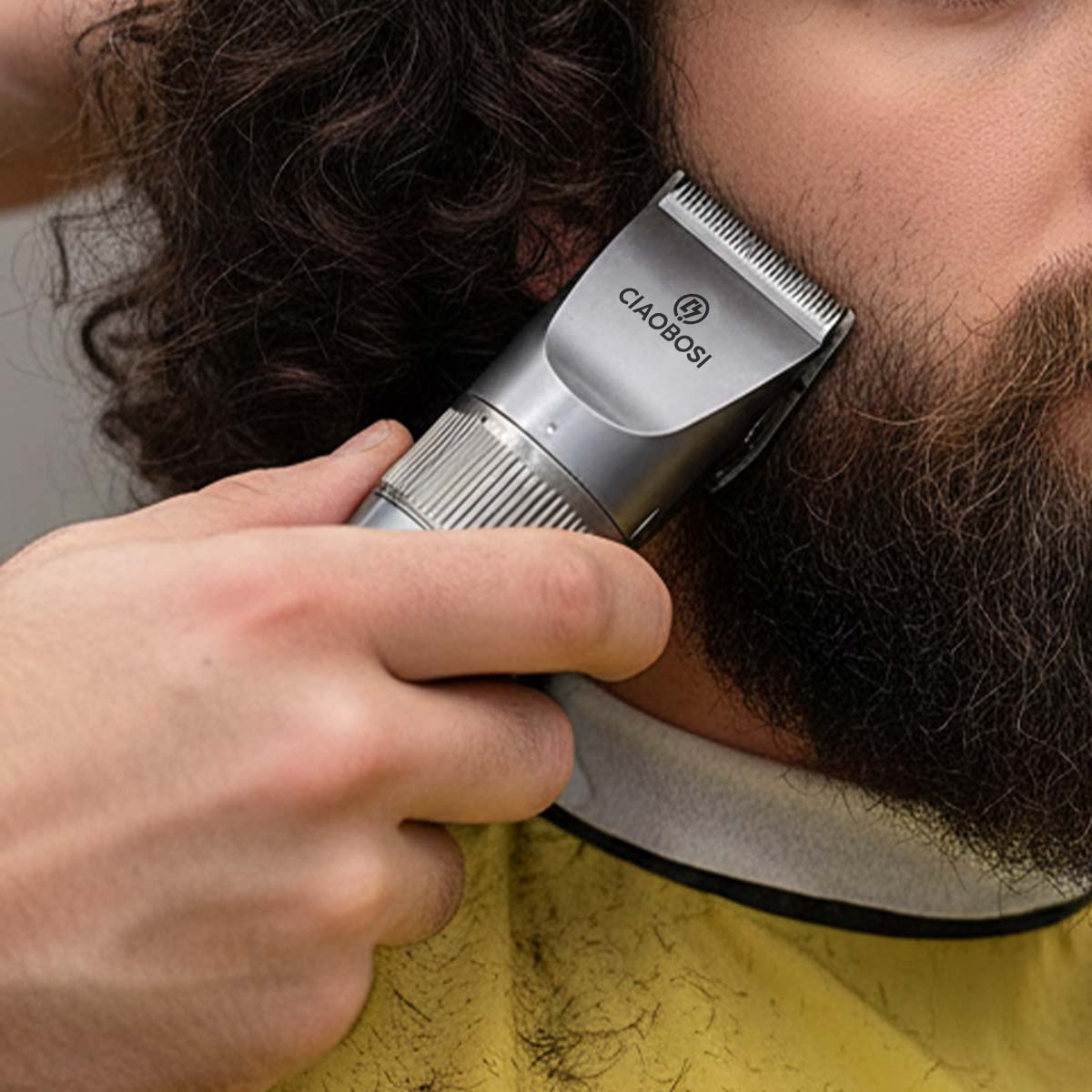 Une personne taille une barbe avec une tondeuse argentée sur une cape jaune.