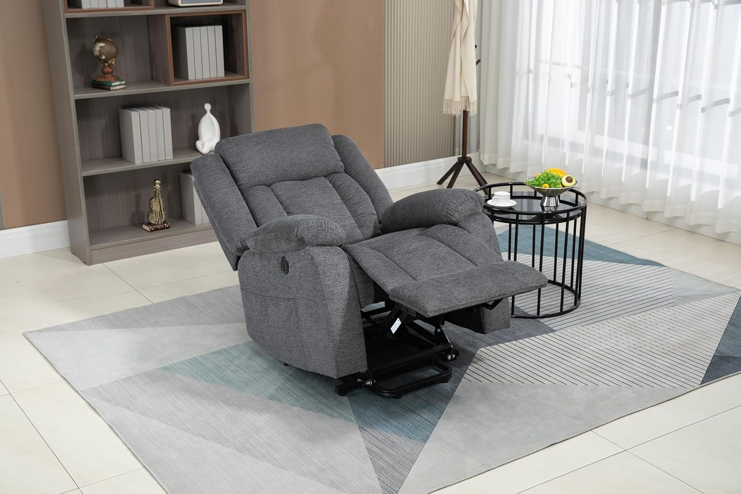 Un sillón reclinable gris con reposapiés extendido está en una habitación con una estantería y una mesa redonda.