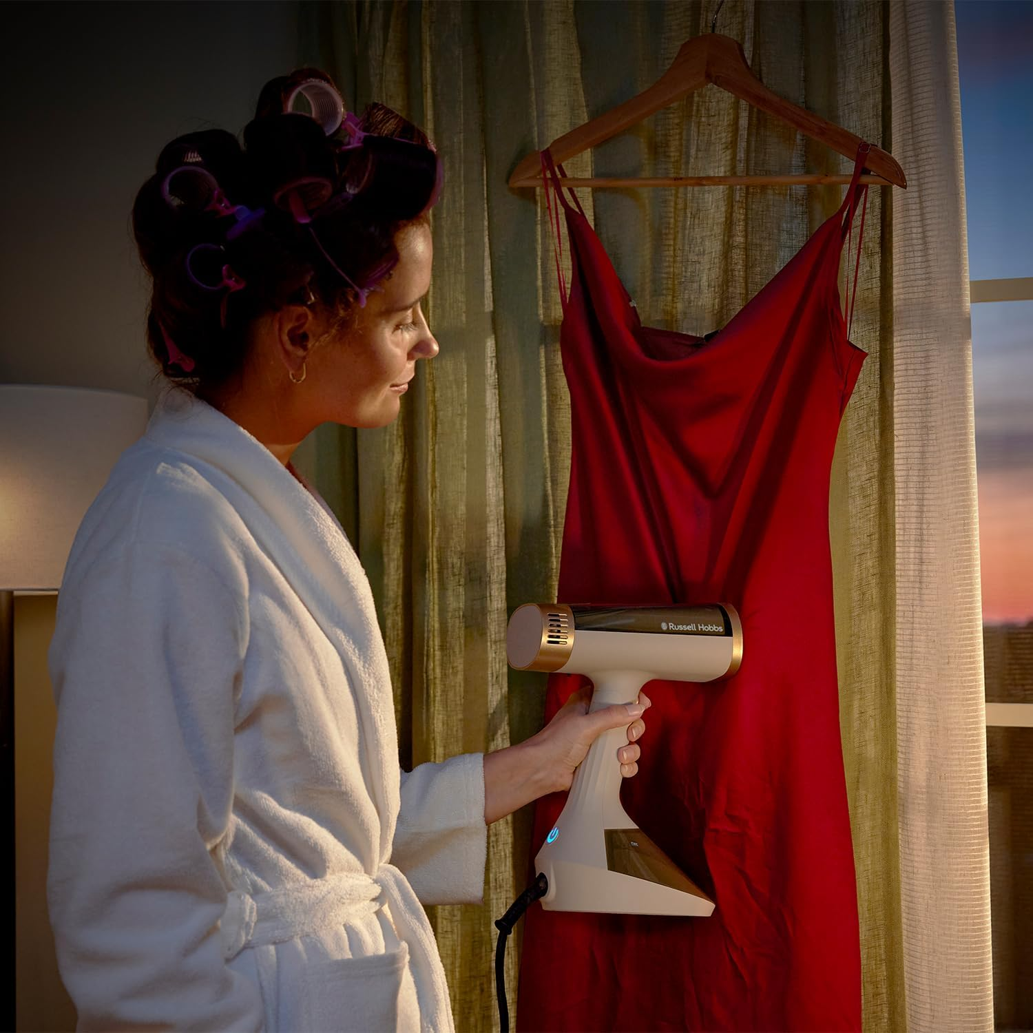 Frau mit Haarwicklern glättet rotes Kleid mit Dampfglätter vor dem Fenster.