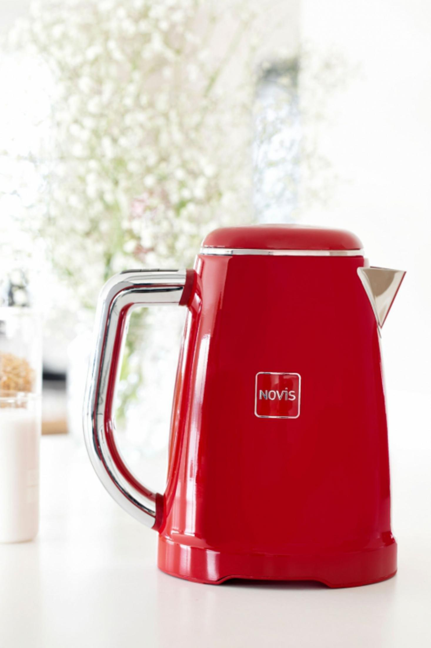 Ein roter Wasserkocher mit Chromgriff steht auf einer weißen Oberfläche, daneben ein Glas.