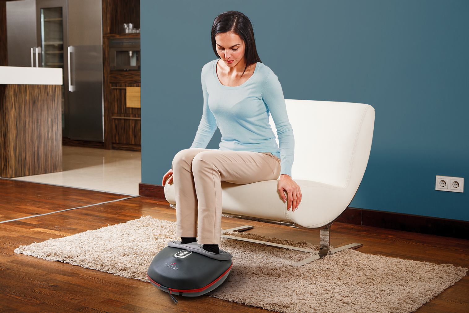 Kobieta siedzi na białym fotelu, stopy w masażerze na dywanie.