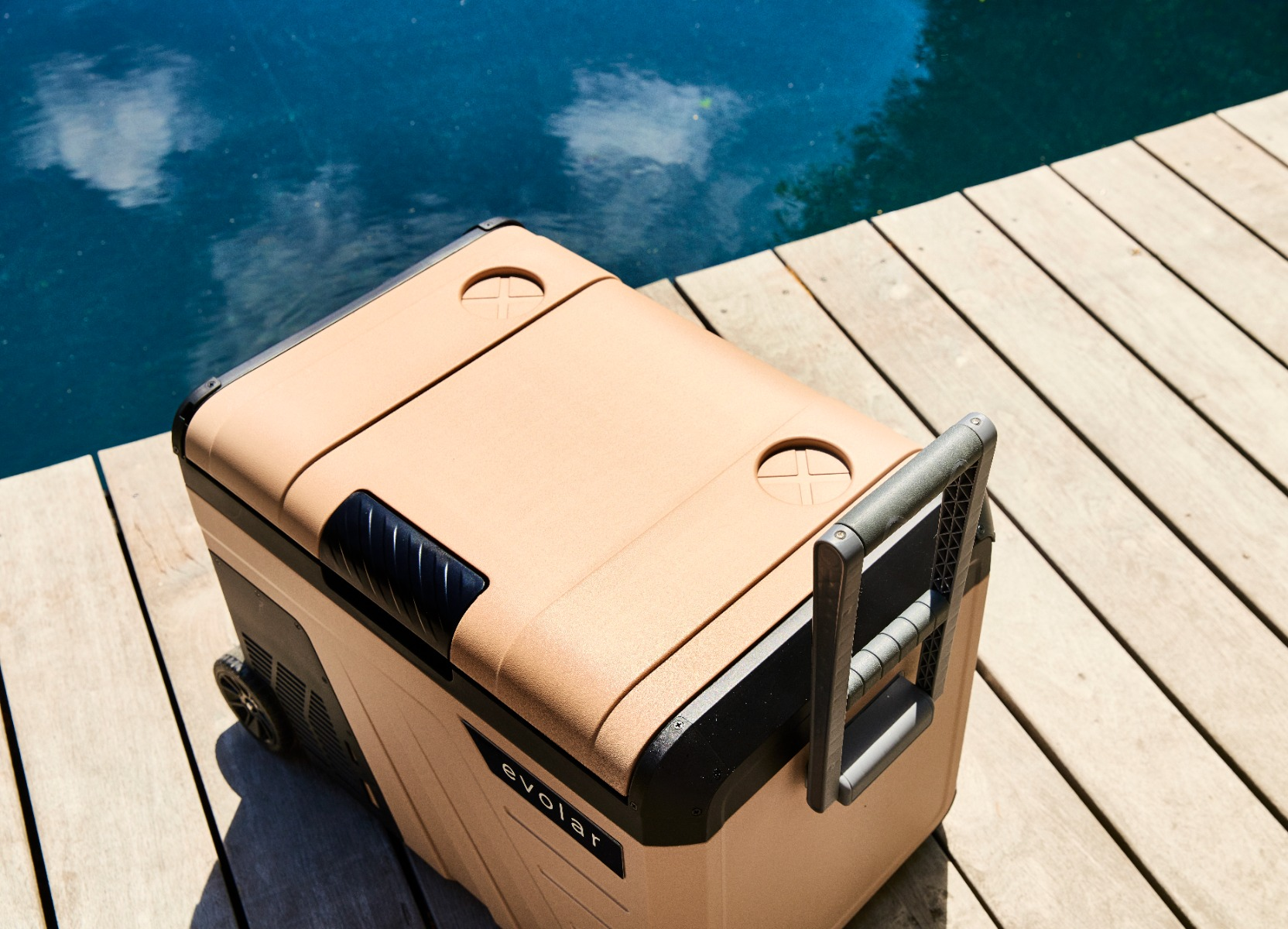 Una borsa frigo beige con ruote si trova su un ponte di legno vicino all'acqua blu.