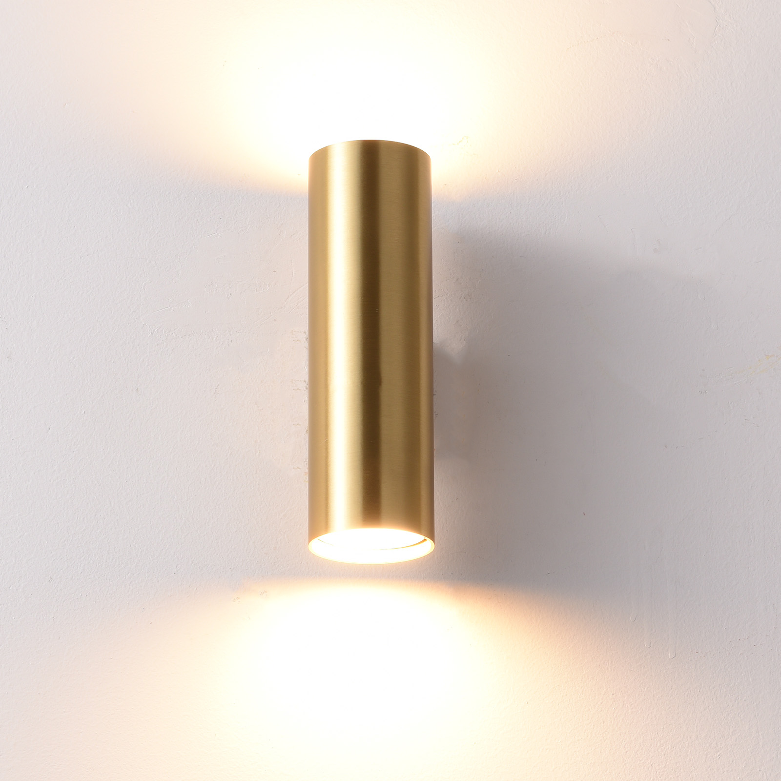 Una lámpara de pared cilíndrica dorada ilumina una pared blanca.