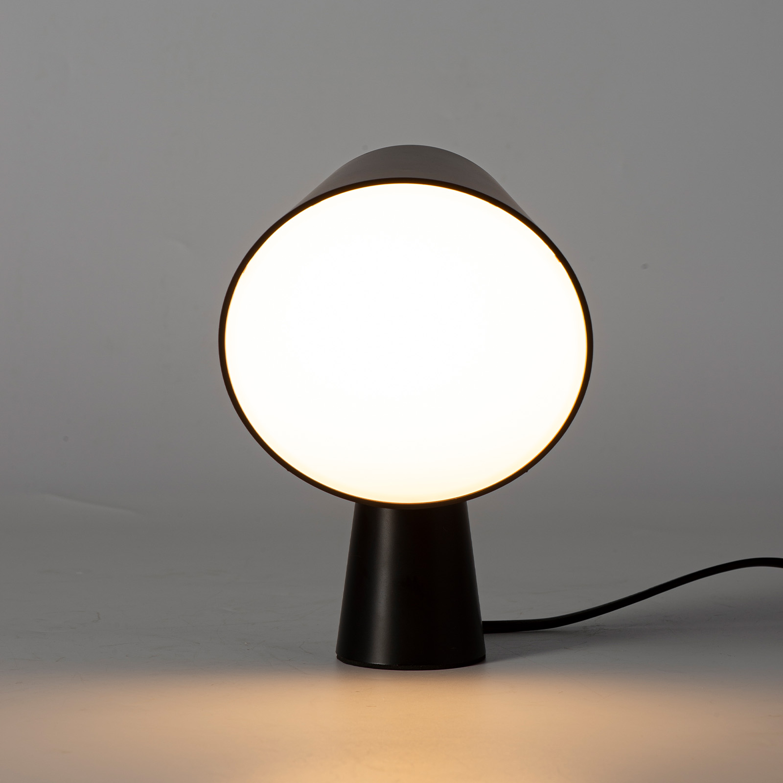 Lámpara de mesa negra con pantalla redonda e iluminada sobre fondo gris.