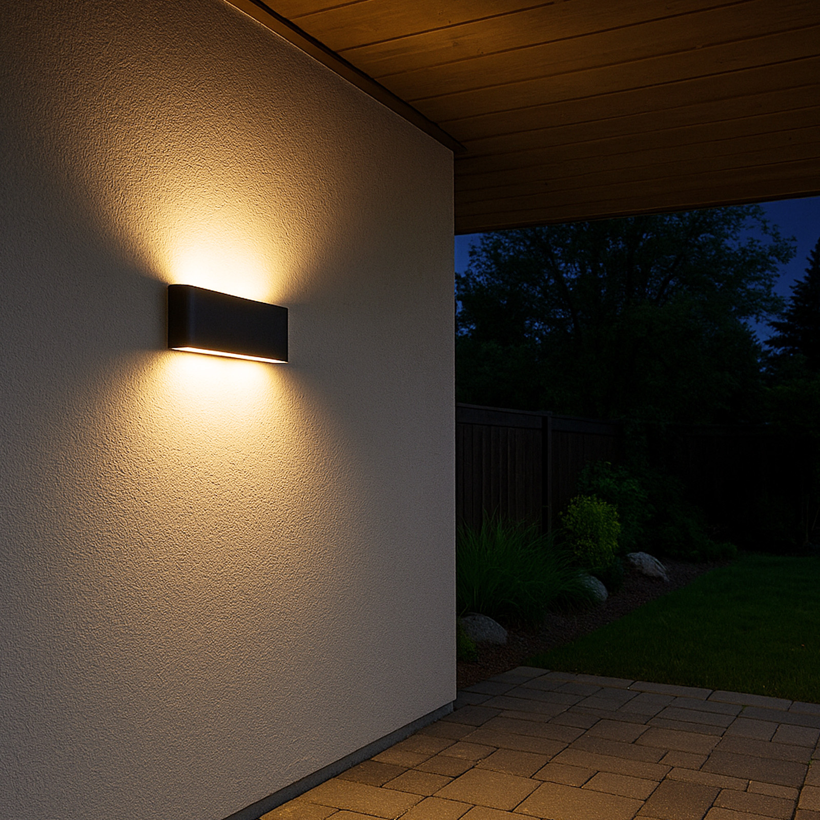 Una lámpara de pared ilumina una pared blanca, junto a un jardín oscuro.