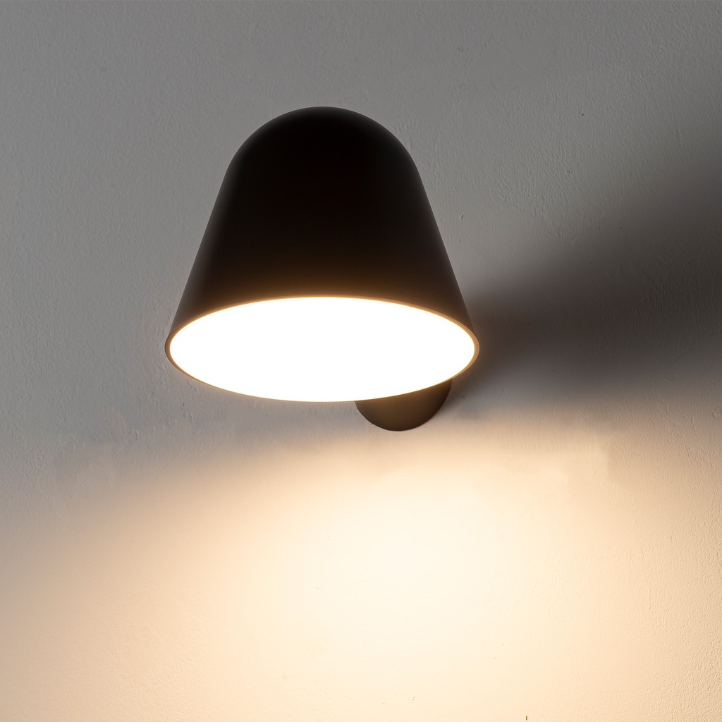 Una lámpara de pared de metal negra con pantalla cónica emite luz brillante en una pared blanca.