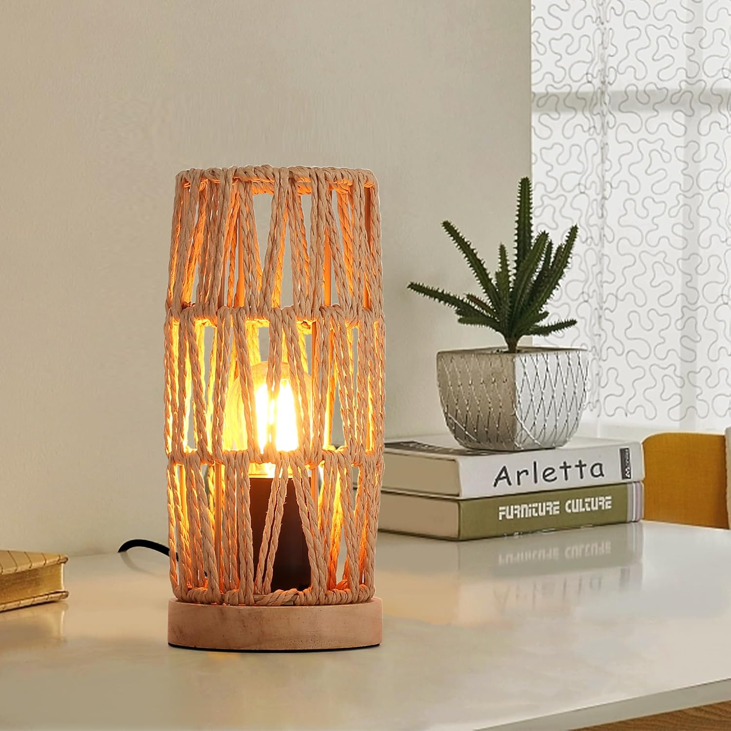 Una lámpara de mesa tejida con bombilla se sienta sobre una mesa junto a libros y una planta.