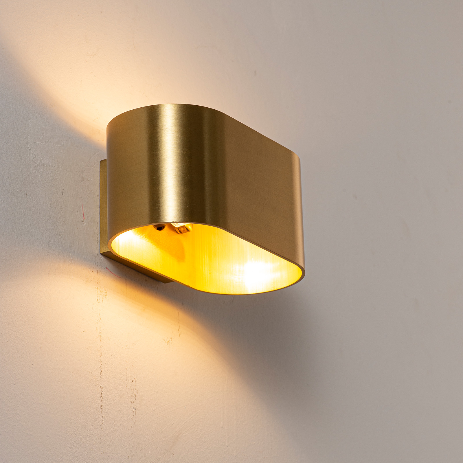 Una lámpara de pared dorada curvada ilumina una pared blanca con luz cálida.
