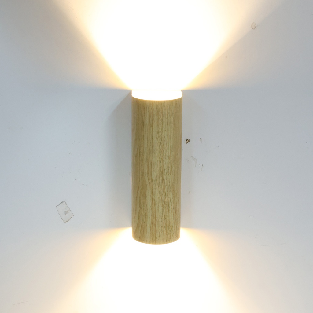 Una lámpara de pared cilíndrica de aspecto de madera ilumina una pared blanca hacia arriba y abajo.
