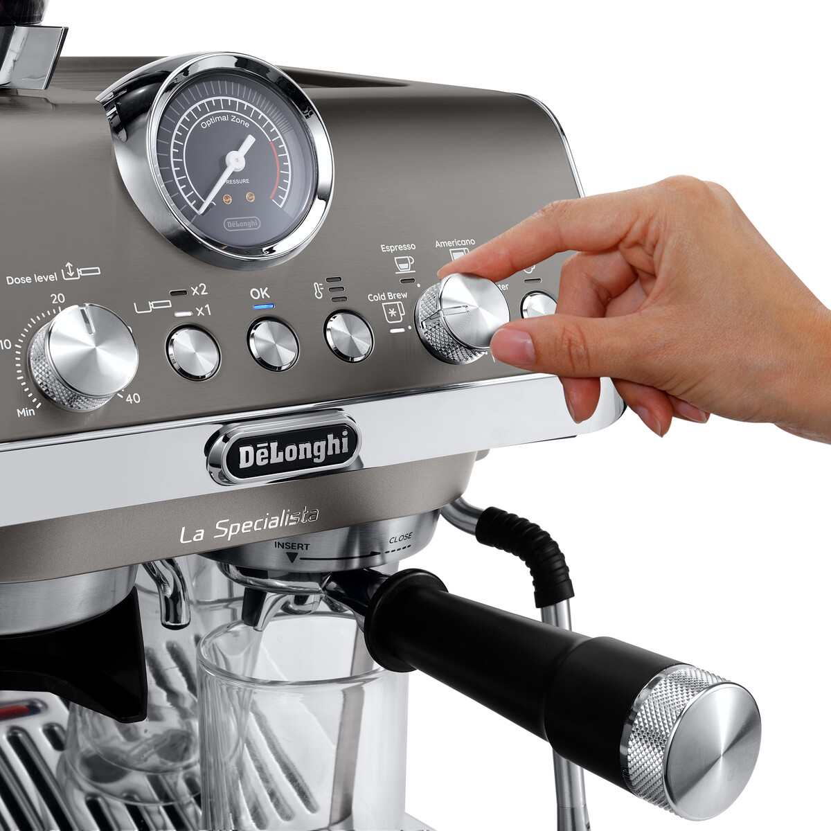 Eine Hand dreht am Knopf einer silbernen DeLonghi Espressomaschine neben einem Glas.