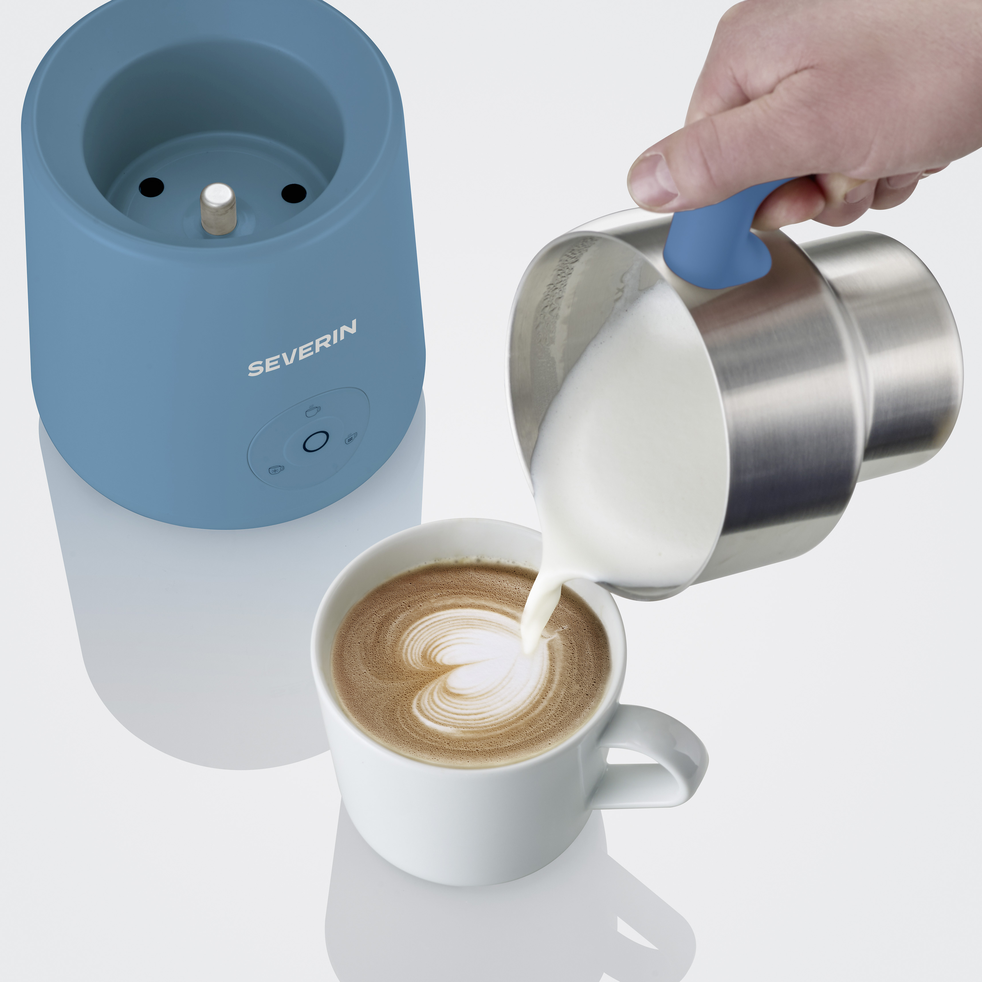 Milchschaum wird in eine Kaffeetasse gegossen, neben einem blauen Milchaufschäumer.