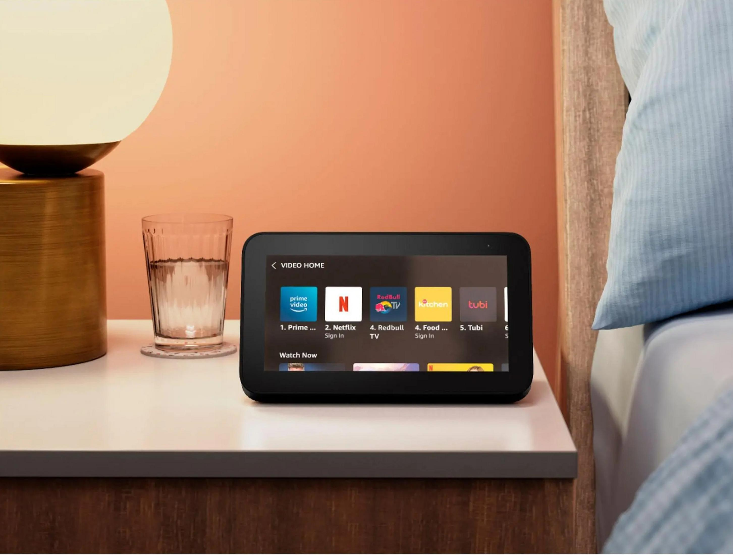 A nightstand with a lamp, glass of water, and a smart display showing video apps.