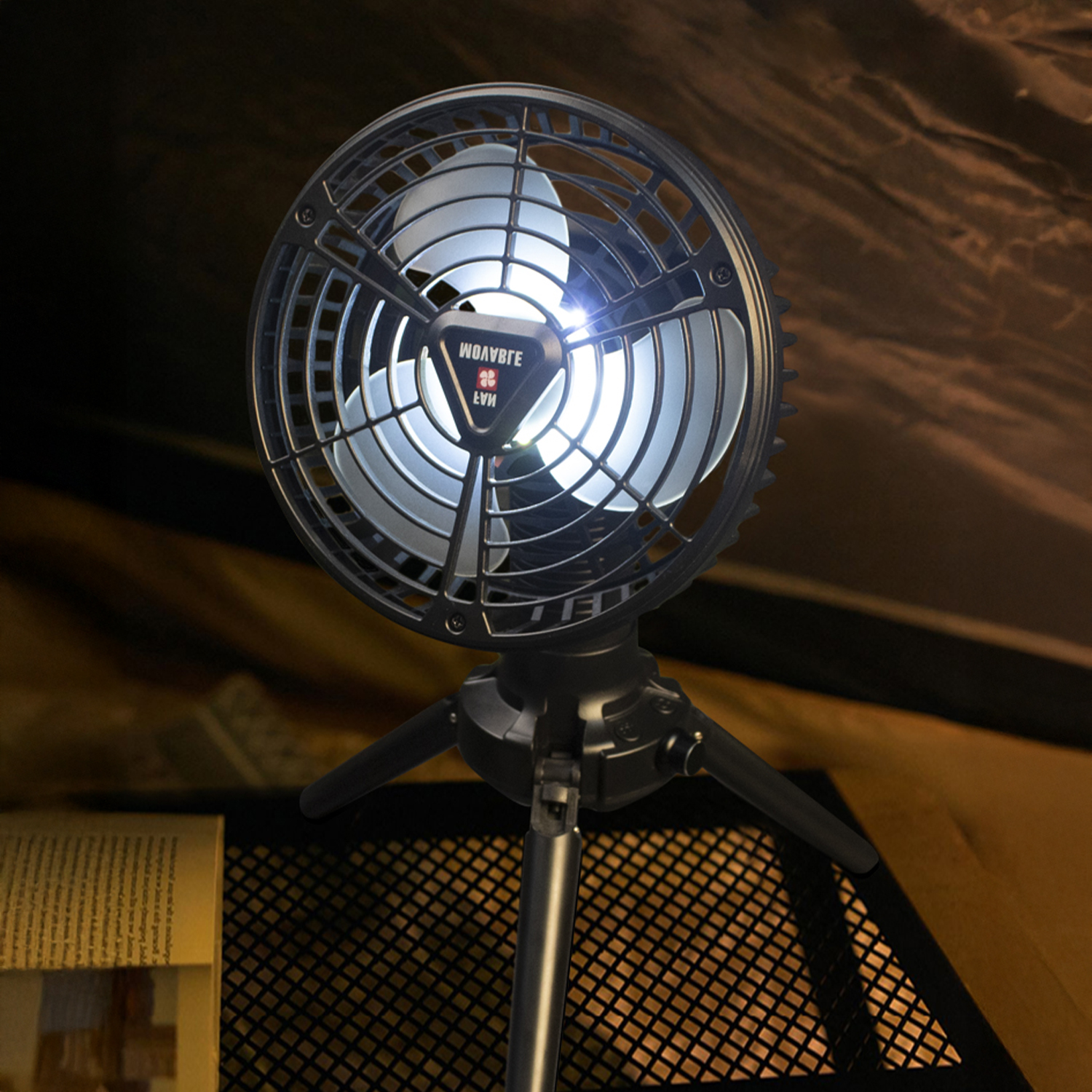 Un ventilatore portatile nero con treppiede è illuminato e posto su un tavolo a rete.