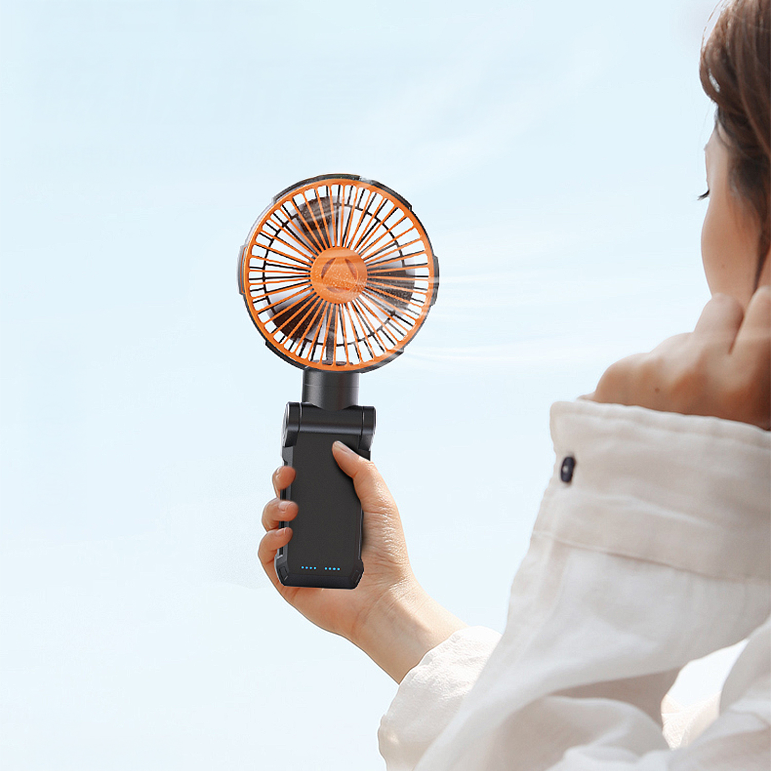 Una mano tiene un ventilatore portatile con pale arancioni contro un cielo blu.