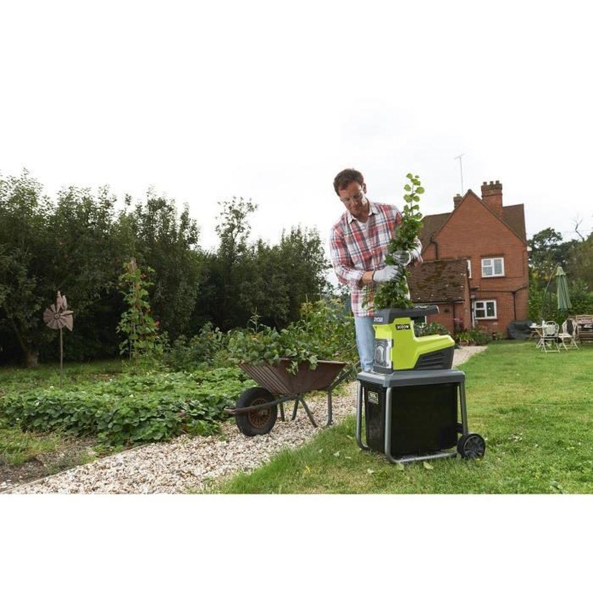 Mężczyzna wkłada roślinę do rozdrabniacza w ogrodzie, dom w tle.