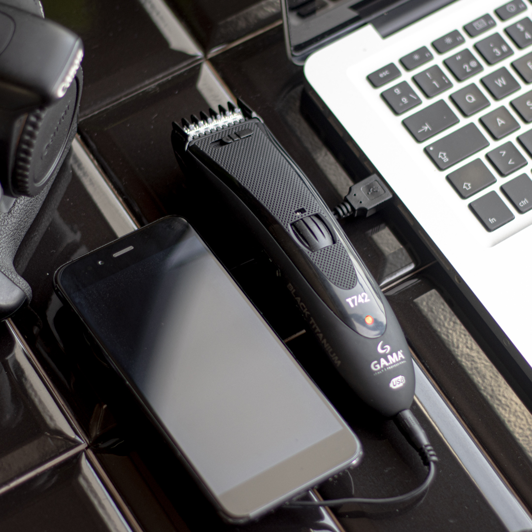Un tagliacapelli nero si carica via USB accanto a uno smartphone e un laptop.