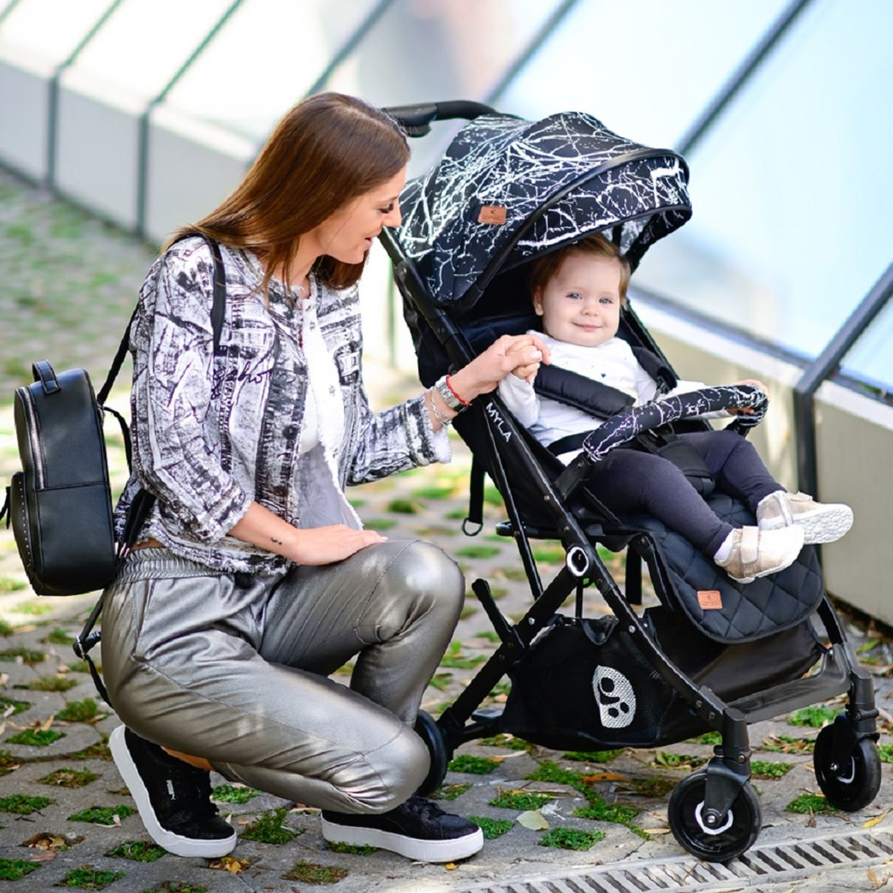 Eine Frau hockt neben einem Kinderwagen mit einem Baby darin. Schwarz-weiße Jacke.