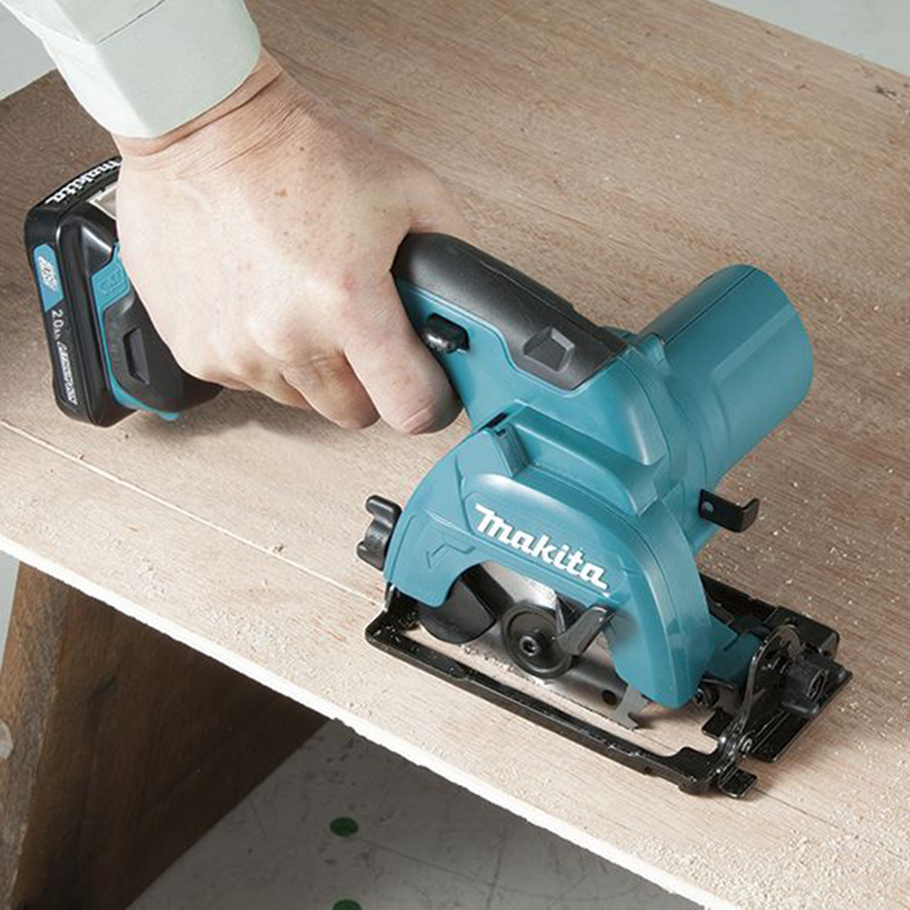 Eine Hand hält eine türkisfarbene Makita-Kreissäge über eine Holzoberfläche.