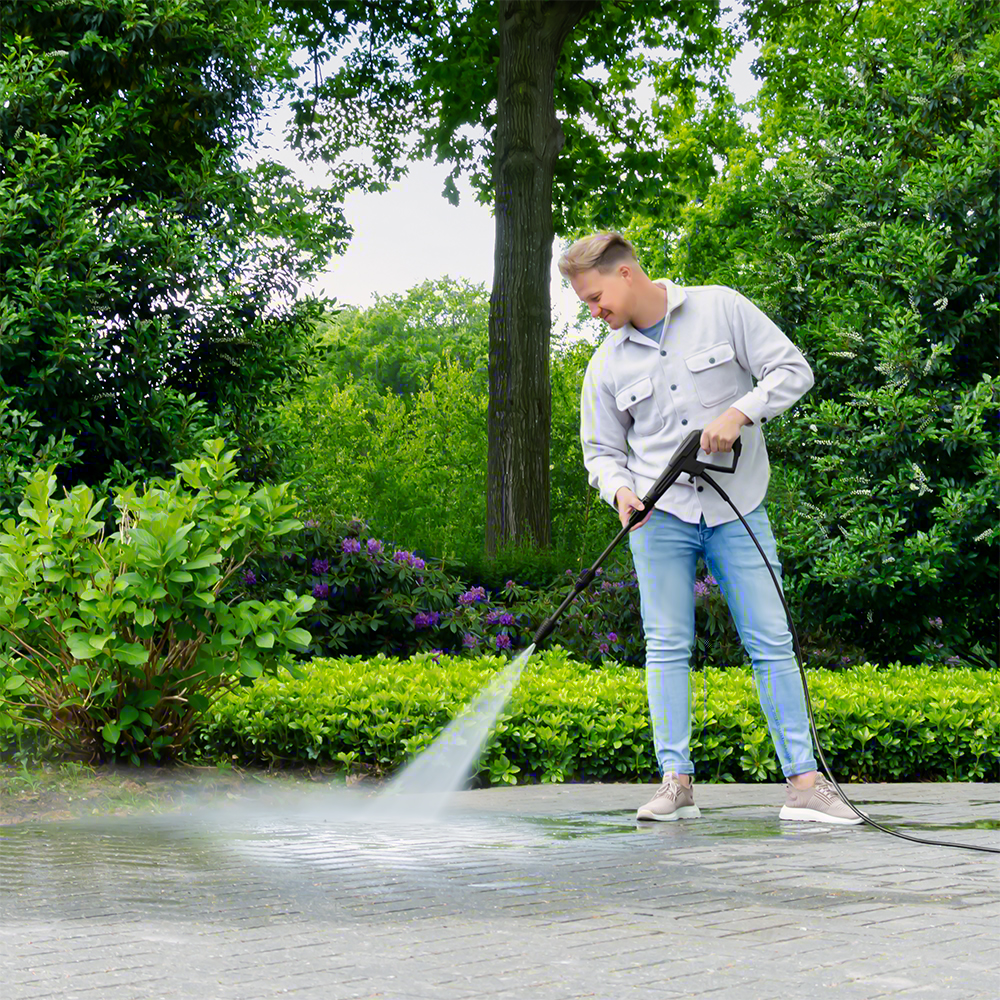 Man reinigt betegeld pad in tuin met hogedrukreiniger. Groene struiken en bomen.