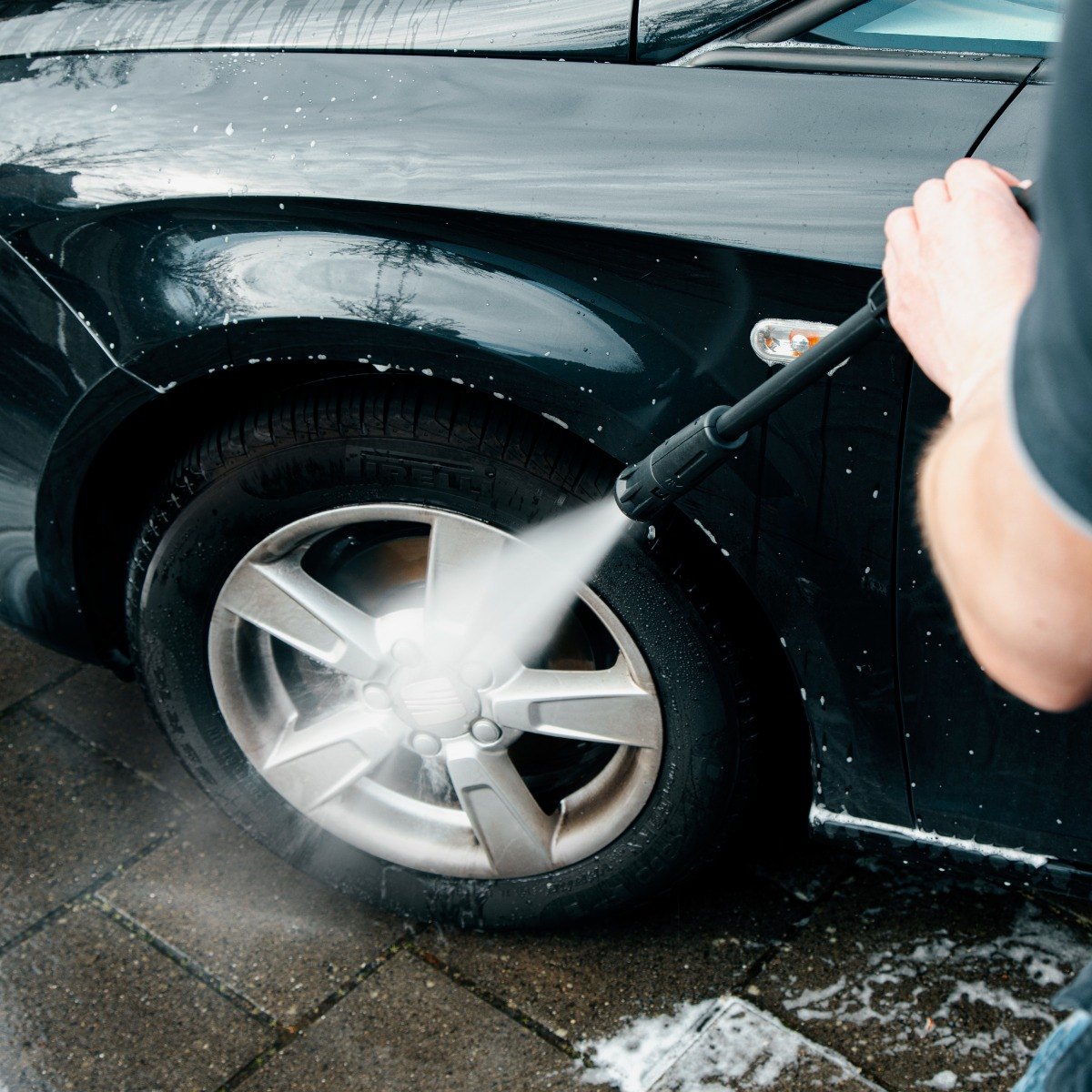 Persoon wast auto met hogedrukreiniger, water spuit op wiel en carrosserie.