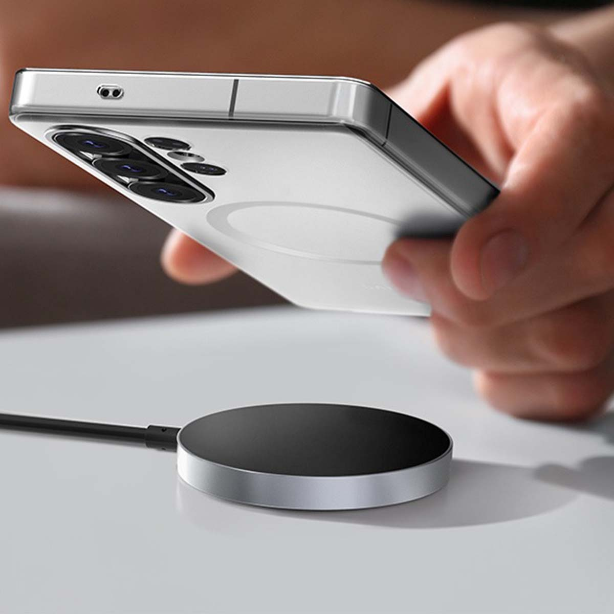 Eine Hand hält ein silbernes Smartphone über eine runde schwarze Ladestation mit silbernem Rand.