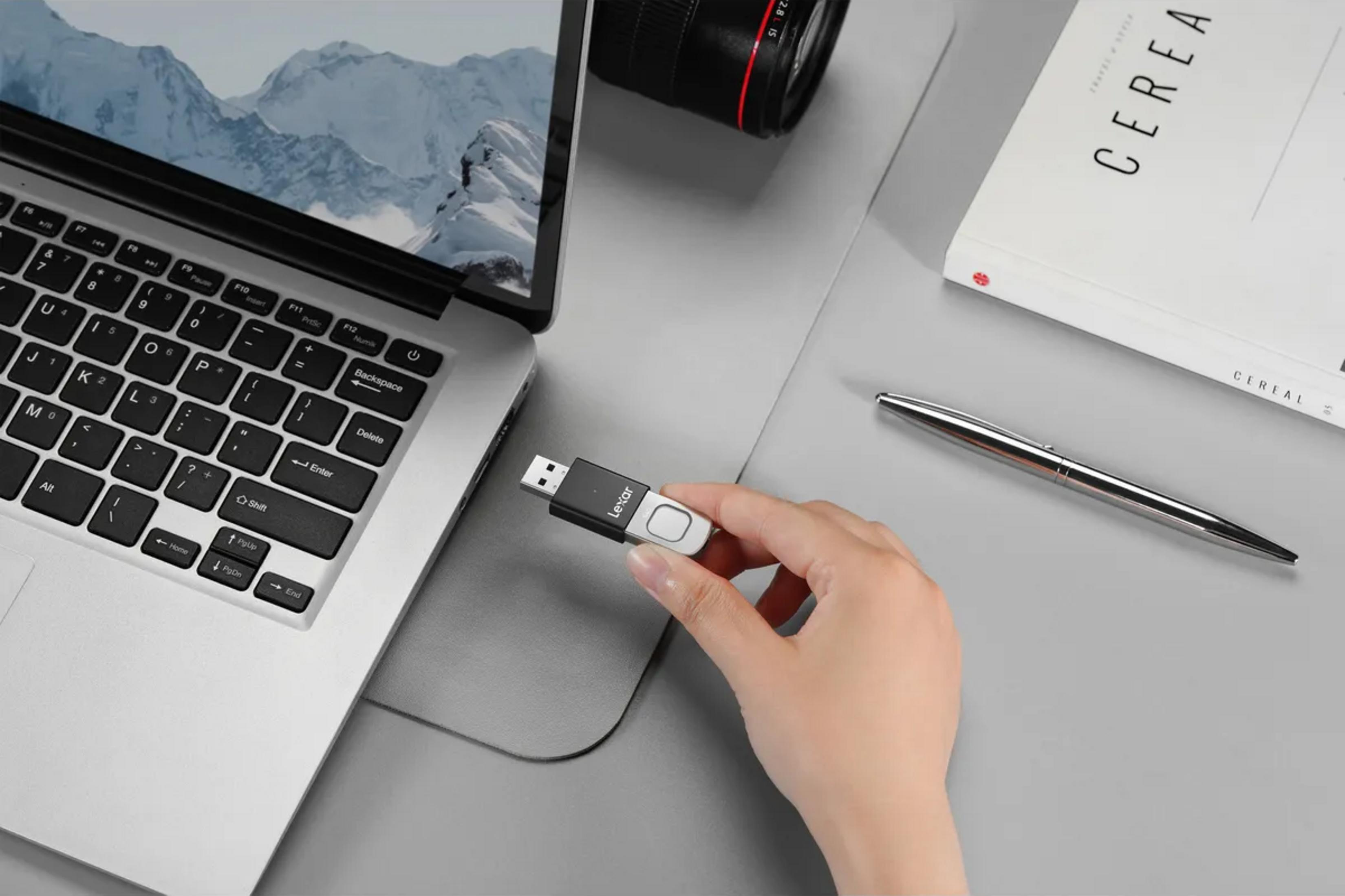 Eine Hand hält einen USB-Stick neben einem Laptop, einem Buch und einem Stift.