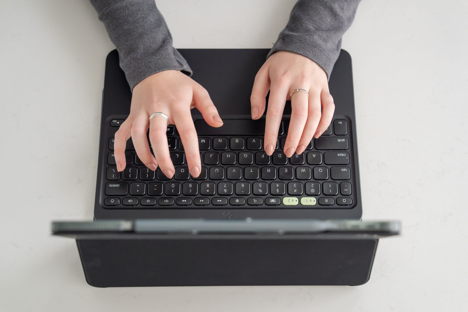 Hände tippen auf einer schwarzen Tastatur, tragen graue Ärmel und Ringe.