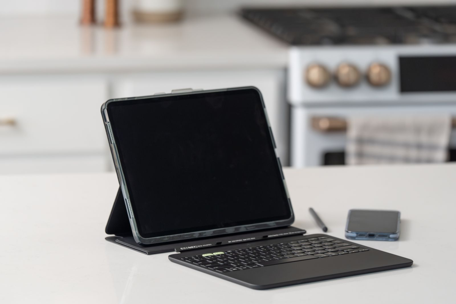 Ein Tablet mit Tastatur und Stift auf einer weißen Oberfläche, Küche im Hintergrund.