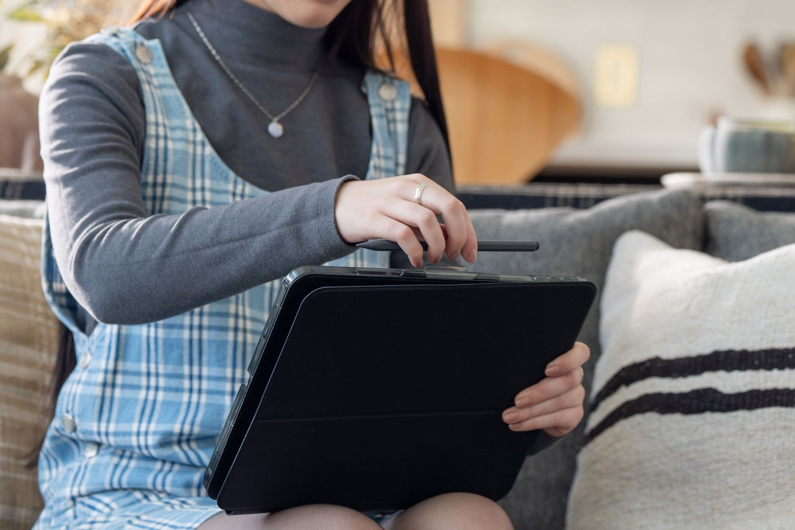 Eine Person in blau kariertem Kleid und grauem Rollkragenpullover hält ein schwarzes Tablet und Stift.