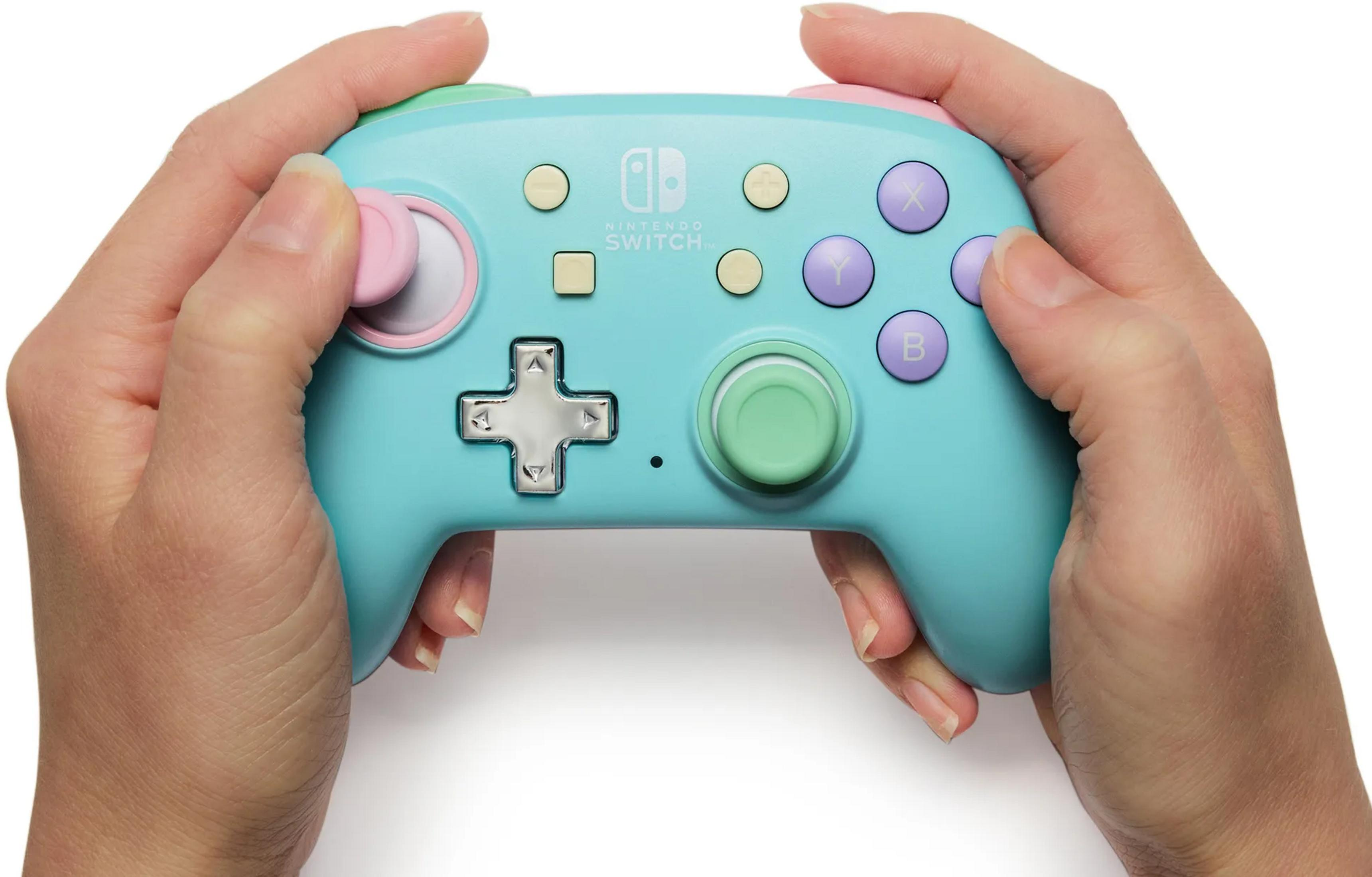 Mains tenant une manette Nintendo Switch turquoise avec des accents roses et verts.