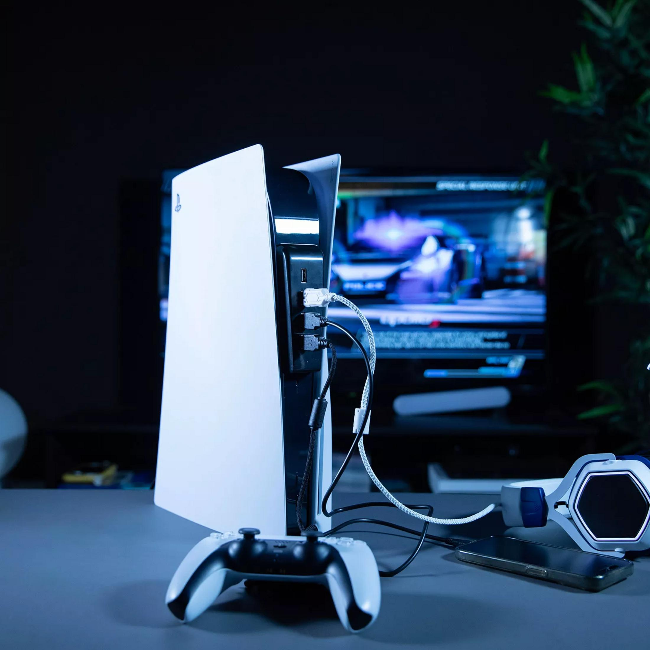 Console PS5 con controller, cuffie e telefono su una scrivania con TV sullo sfondo.