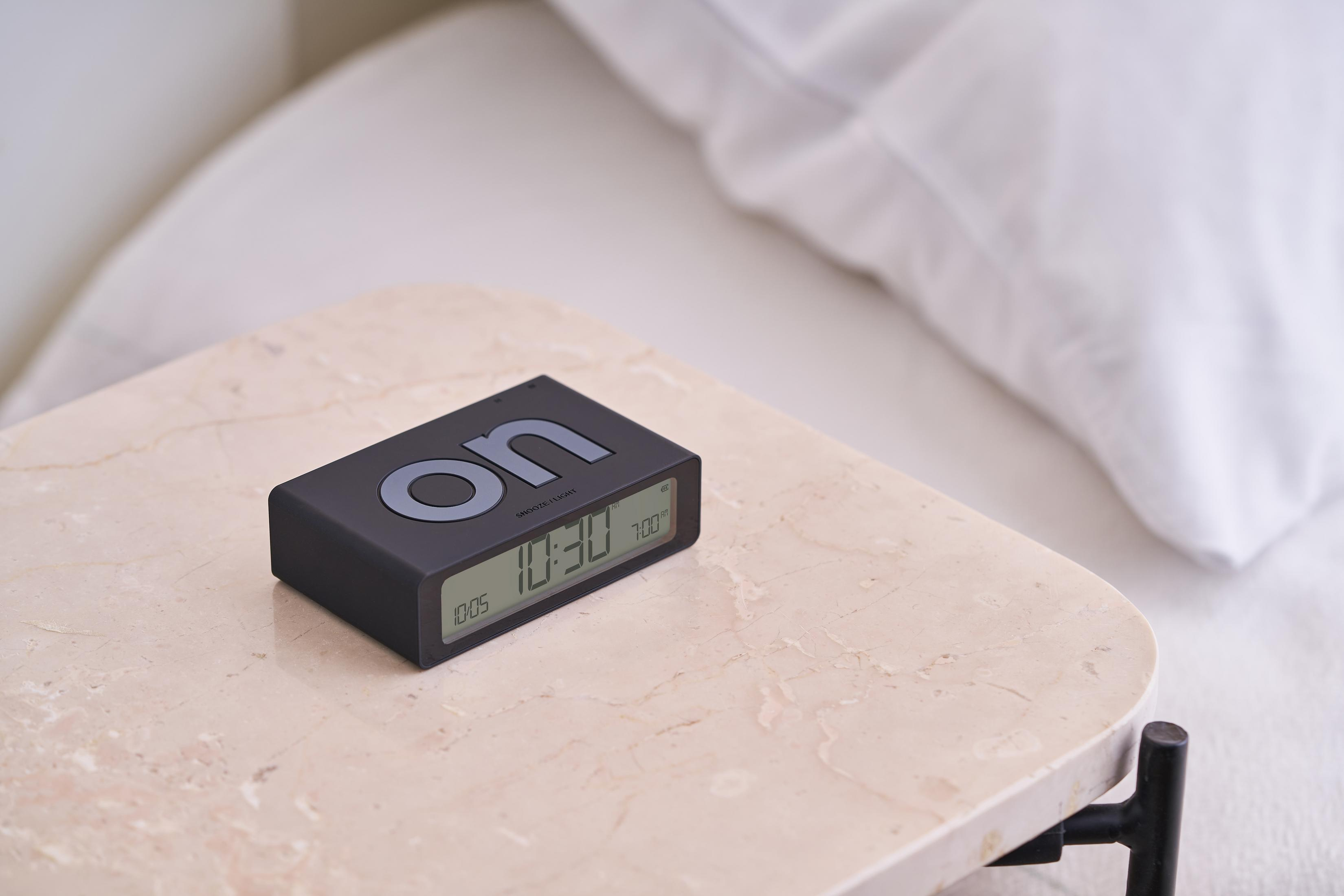 Schwarzer Digitalwecker auf Marmortisch neben einem Bett mit weißer Bettwäsche.