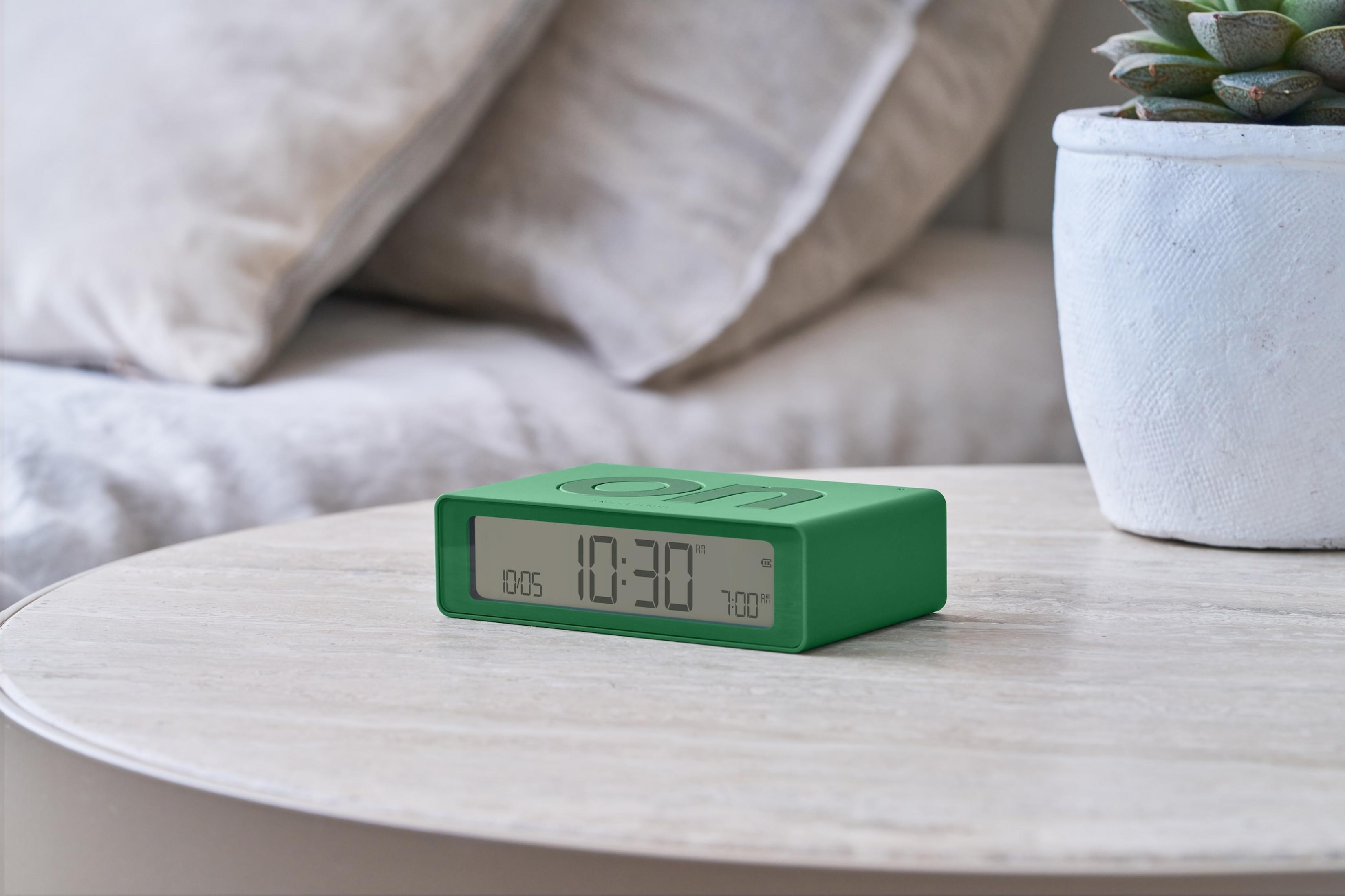 Réveil numérique vert avec deux alarmes sur une table de nuit près des oreillers.