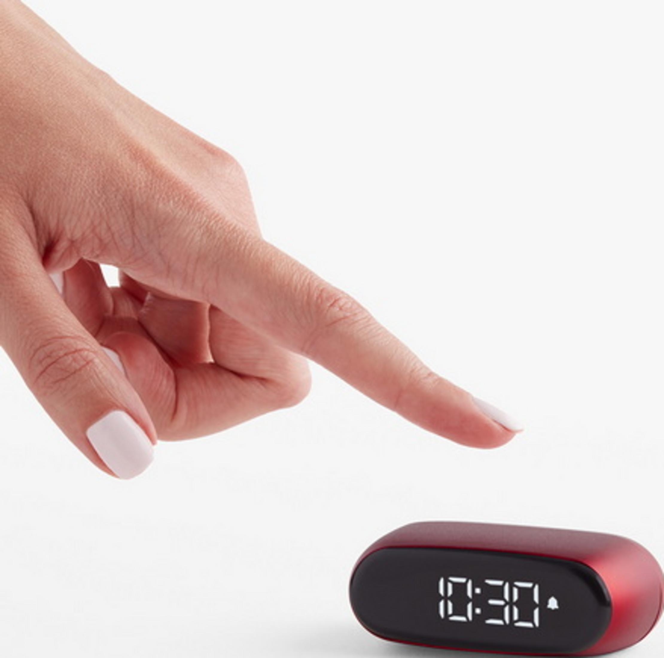Eine Hand mit weiß lackierten Nägeln berührt einen roten Digitalwecker, der 10:30 anzeigt.