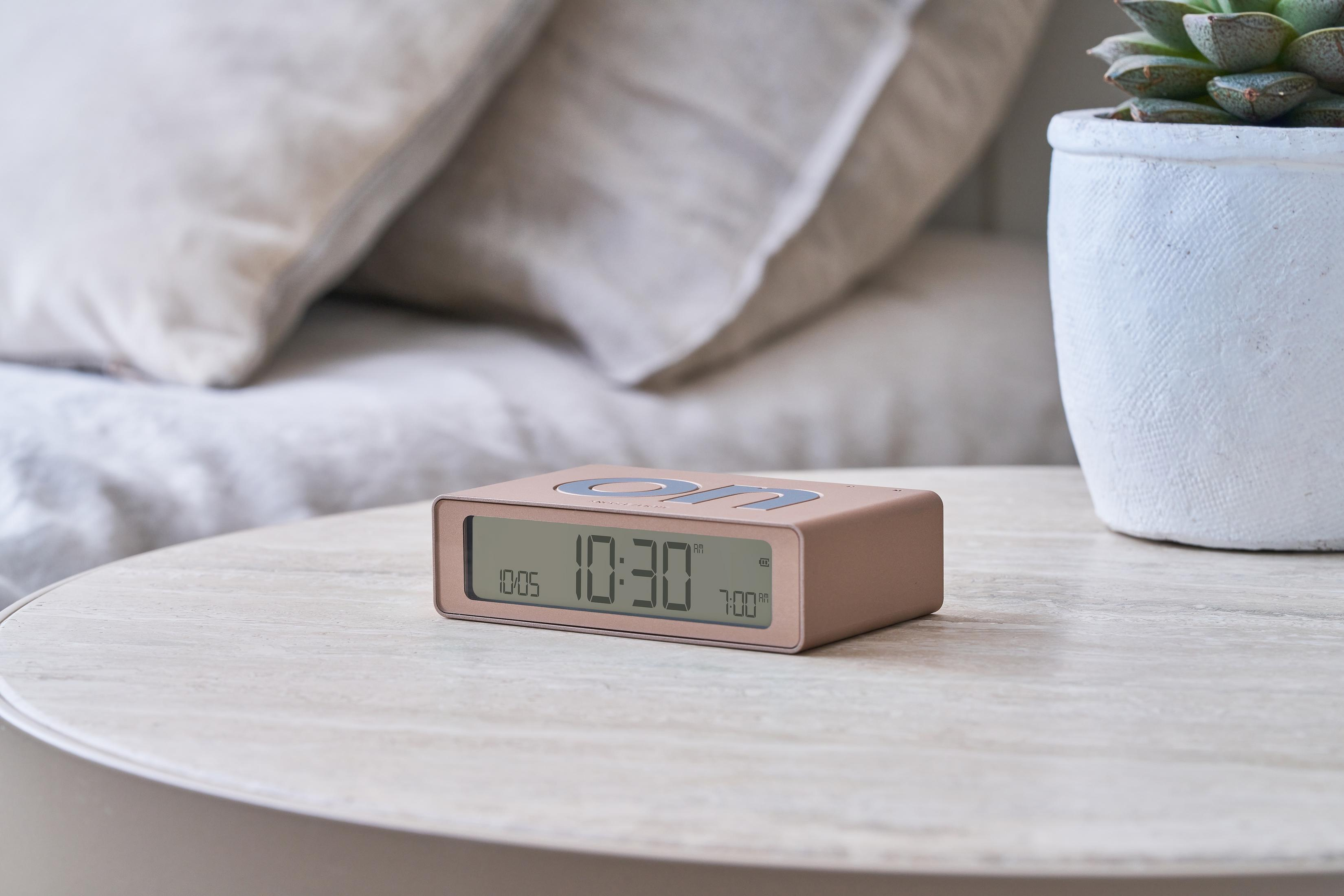 Ein roségoldener Digitalwecker zeigt 10:30 Uhr auf einem Nachttisch neben Kissen.