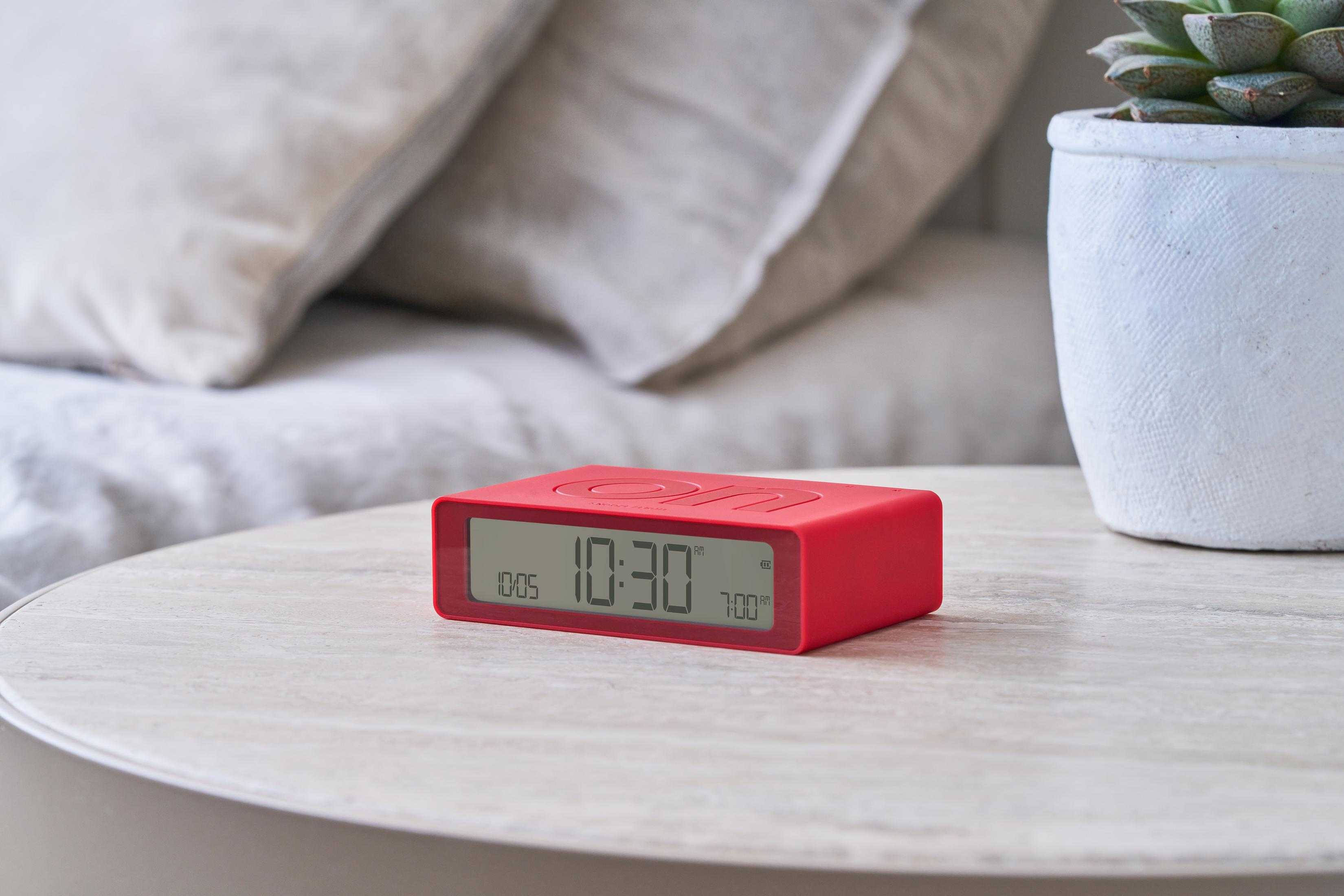 Ein roter Digitalwecker auf einem Nachttisch mit Kissen und Pflanze.