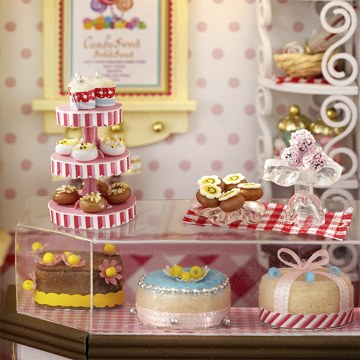 Negozio di caramelle in miniatura con cupcake, ciambelle e torte esposte dietro vetro su tovaglie a scacchi.
