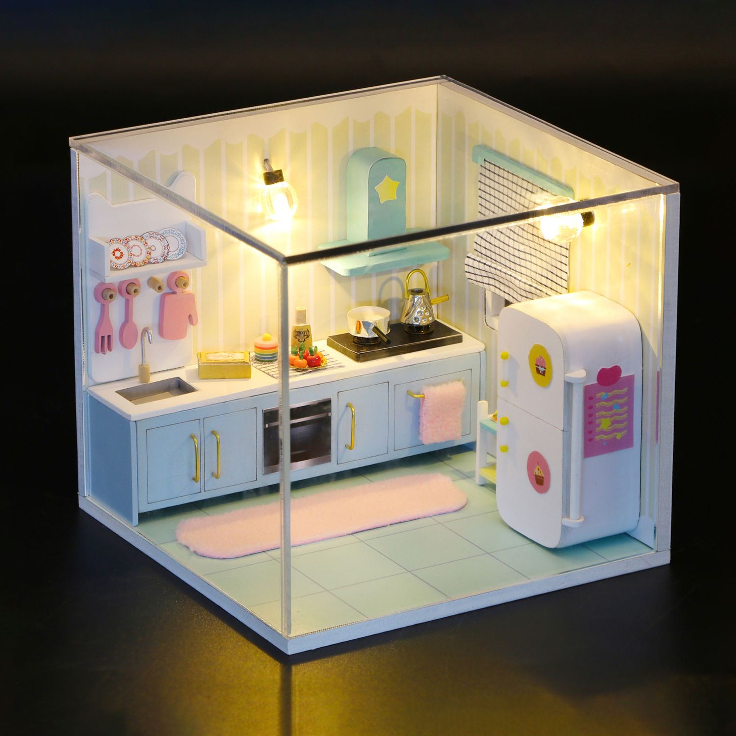 Una cucina in miniatura con mobili blu, frigorifero bianco e accenti rosa. Le luci sono accese.