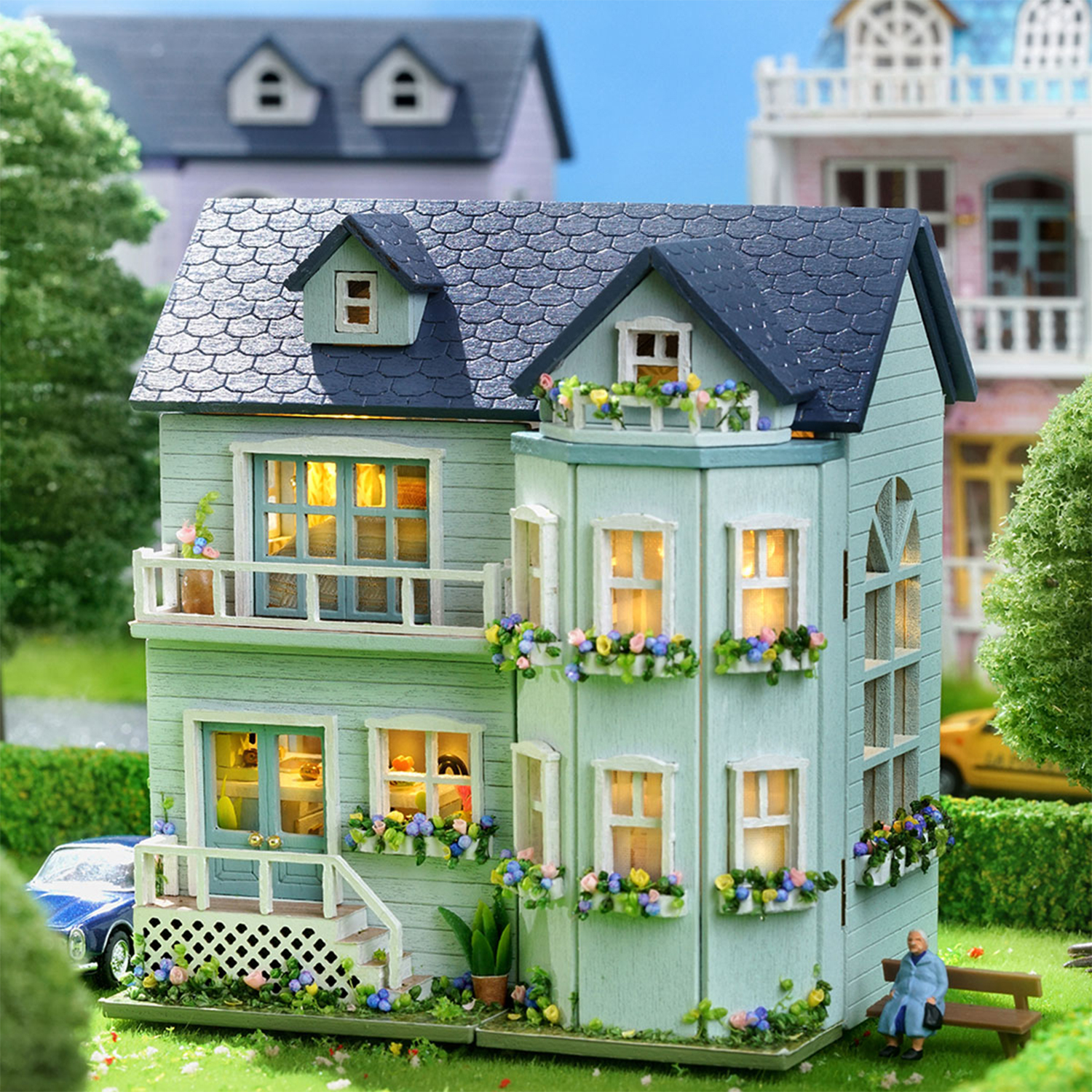Casa in miniatura verde menta con tetto blu, balconi, fioriere e figura su panchina.